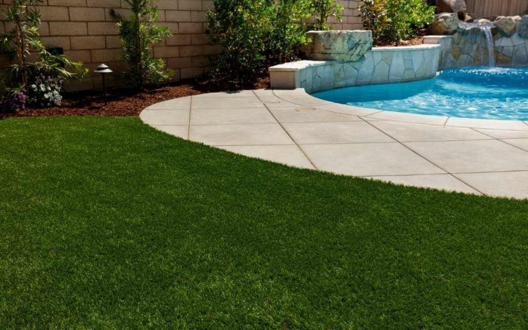 A backyard with a swimming pool and a waterfall feature, surrounded by stone tiles. The pool area is bordered by lush artificial turf. In the background, there is a brick wall and various plants and bushes, showcasing exceptional landscaping typical of Houston, TX.